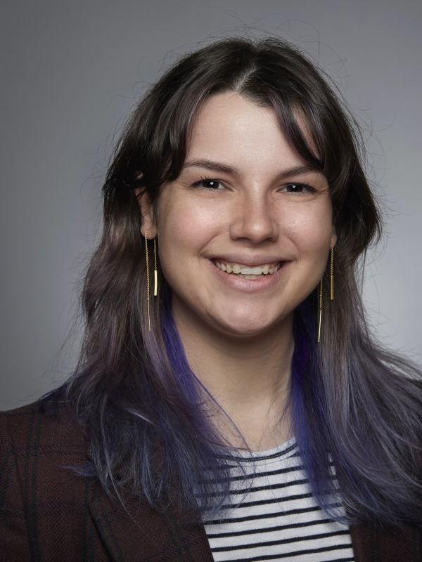 katie smiles at the camera. She has purple and blue hair, is wearing gold drop earrings, and is wearing a black and white striped shirt with a maroon blazer