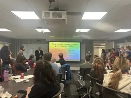 many people from the department, sitting and standing, listening to the department chair speak